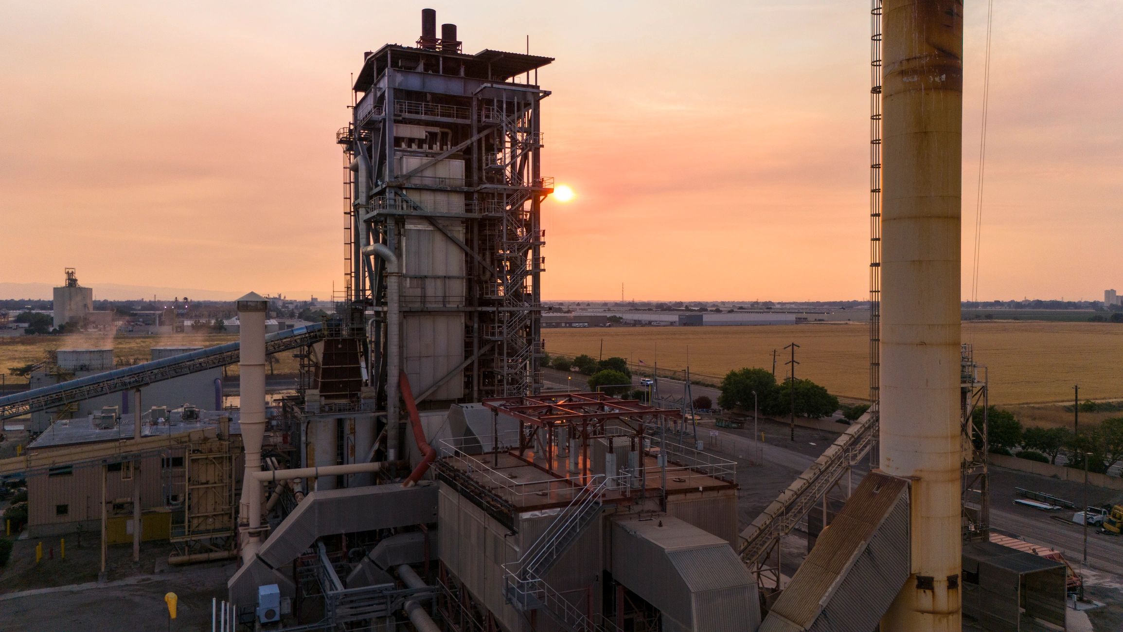 Biomass power plant tower standing tall against a smoky sunset sky. Industrial facility utilizing agricultural waste in California.